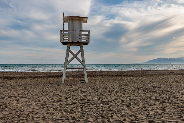 Lifeguard tower stands on a pristine sandy beach under a clear blue sky, facing the calm, vibrant blue ocean. Minimalistic, summery, and tranquil atmosphere perfect for relaxation or coastal themes