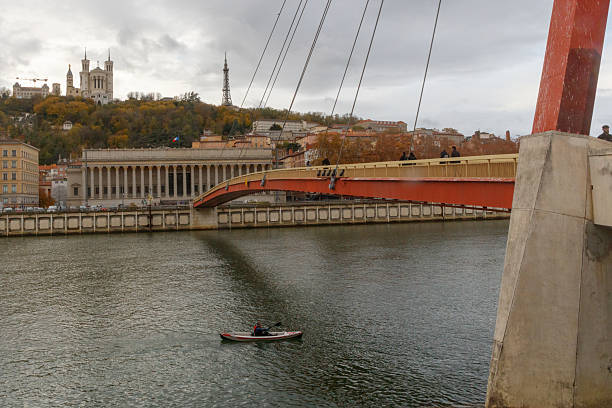 Image of the Pont de la Feuill?e and the Basilica of Notre-Dame de Fourvi?re in Lyon, France, captured from across the Sa?ne River. Several pedestrians can be seen on the bridge, with the basilica and the metallic tower of Fourvi?re visible on the hill in the background, under a cloudy sky.
