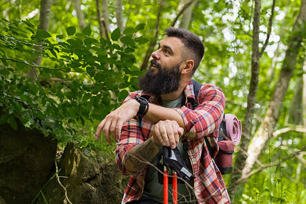 With both hands resting on his poles, a bearded hiker leans on a mossy rock, gazing around the peaceful woodland, clearly enjoying the moment.