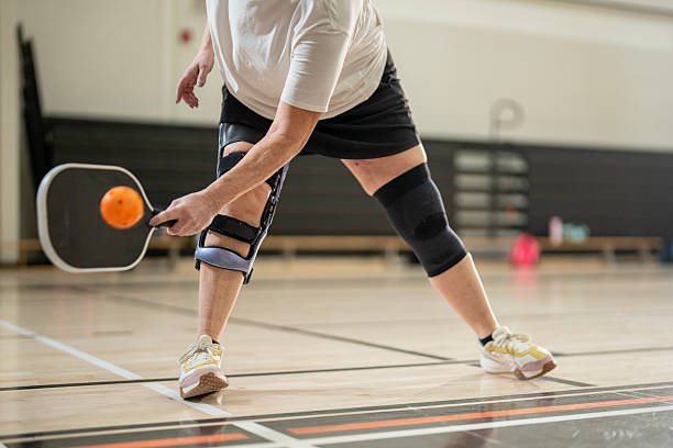 An individual engaged in a pickleball match indoors, showcasing their active pose while reaching for the ball, equipped with knee braces for support. The setting is bright, focused, and sporty.
