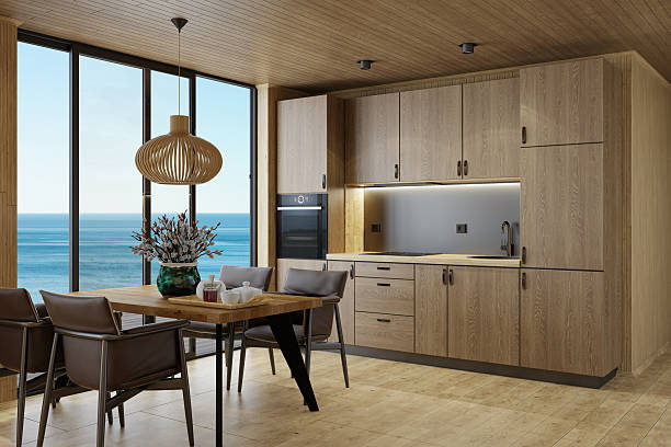 Modern Scandinavian log cabin kitchen entirely crafted from natural oak wood, featuring eco-conscious design and warm textures. The space includes oak hardwood flooring, oak wall and ceiling paneling, and oak kitchen cabinets with a minimalist aesthetic. The dining area is furnished with an oak wood table and four leather chairs. Large floor-to-ceiling windows and sliding glass doors open to a breathtaking panoramic view of the ocean under a clear sky. Ideal for promoting sustainable living, eco design, architectural concepts, luxury cabins, and real estate marketing.