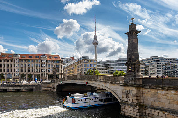 Berlin, Germany, May 26, 2025: Tour boat passes under a stone bridge on the River Spree in Berlin, with the iconic TV Tower and modern buildings in the background