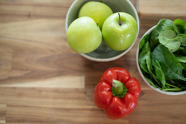 An overhead view of green apples, a red bell pepper, and spinach leaves in bowls on a wooden surface. The arrangement emphasizes freshness, healthy living, and natural ingredients.