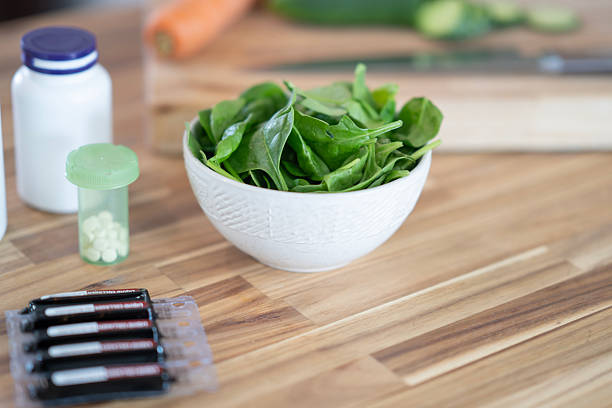 A bowl of fresh spinach is accompanied by vitamins and supplements on a wooden counter, representing healthy living.