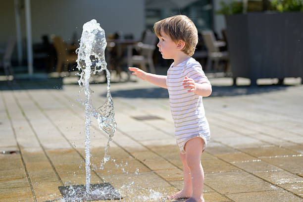 A young child stands barefoot in front of a ground-level water fountain, reaching out with curiosity.