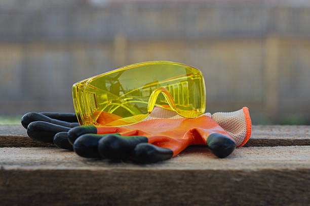 Yellow safety goggles and orange gloves rest on a rustic wooden surface. Concept workplace safety for construction or DIY tasks