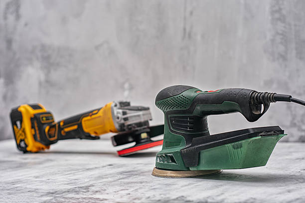 Cordless grinder and a sander rest on a concrete surface against a textured wall. Tools for construction and DIY projects.