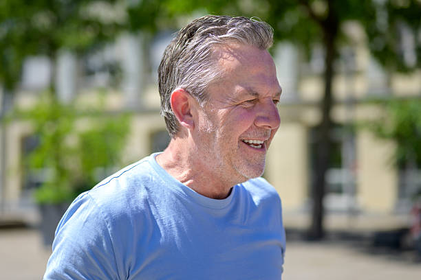 Portrait of a happy elderly man with grey hair and a trimmed beard, wearing a light blue T-shirt, smiling while enjoying a sunny day outside.