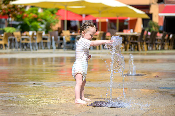 Woman in a white dress enjoys the playful water streams of a fountain at Dubai Creek Harbour with boats docked nearby, capturing a moment of innocent fun under the bright sun.
