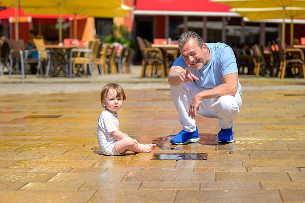 Smiling elderly man squatting beside a young child sitting barefoot on wet pavement, with colorful outdoor café seating and umbrellas in the background.