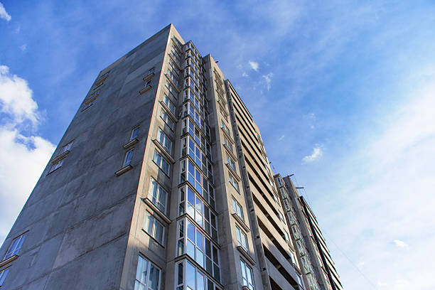 A tall high-rise apartment building with numerous windows rises against a cloudy blue sky. Urban architecture and residential living.