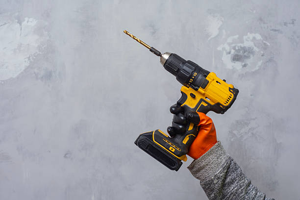 istock Worker in an orange glove holds a cordless drill with a bit, set against a textured gray background. Powerful tool for construction and DIY projects. 2218135715