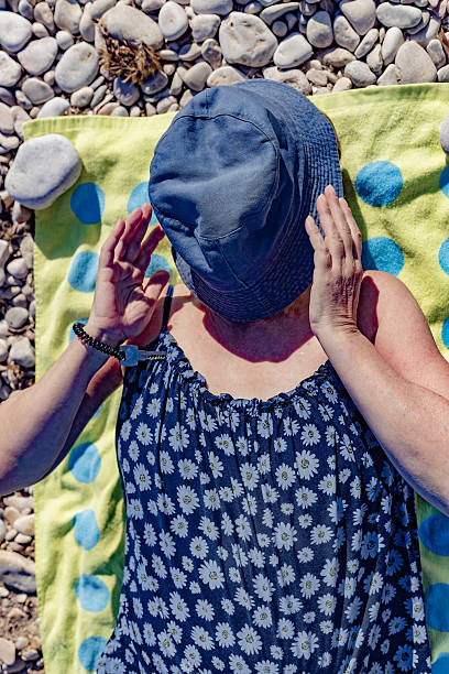 Altea. Alicante. Spain. A woman is lying on a colorful beach towel on a pebble beach, covering her face with a hat, enjoying the summer sun and relaxing during her vacation