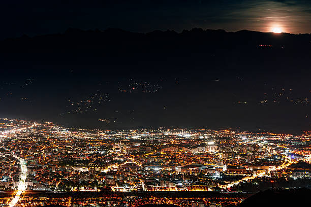 Aerial view of a city illuminated at night with the moon rising over distant mountains. Urban lights create a vibrant atmosphere under the night sky.