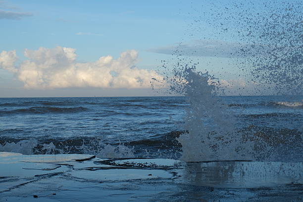 The image captures a tranquil coastal scene where small waves gently roll onto the beach. The deep blue ocean contrasts beautifully with the soft hues of the sky, dotted with white clouds. The calm ambiance and natural lighting make this a peaceful and evocative seaside landscape, perfect for themes of nature, relaxation, and coastal beauty.