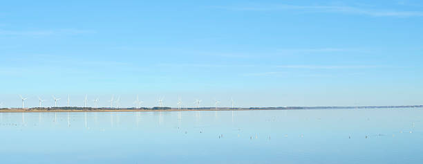 A picturesque view of wind turbines along a serene coastline in Denmark. The calm water reflects the clear blue sky, epitomizing sustainable energy in harmony with nature.