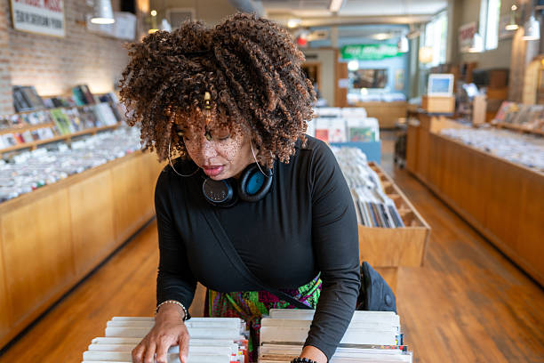 A record store clerk stands behind the counter organizing stacks of vinyl records in a small independent music shop. The interior is packed with albums, posters, and music memorabilia, with bright yellow-and-black bins of records in the foreground and colorful record sleeves covering the walls.
