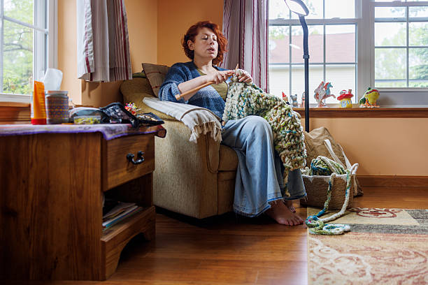 A 47-year-old Caucasian woman with curly hair sits in a gray upholstered chair by large windows, actively knitting a green and white patterned afghan. She holds knitting needles and works with yarn while natural light illuminates her indoor workspace. The cozy living room features hardwood floors, a patterned area rug, and wooden furniture including a side table with craft supplies. Windows reveal green foliage outside in this rural Pennsylvania home setting. The scene captures a quiet moment of focused handcraft work in a comfortable domestic environment.