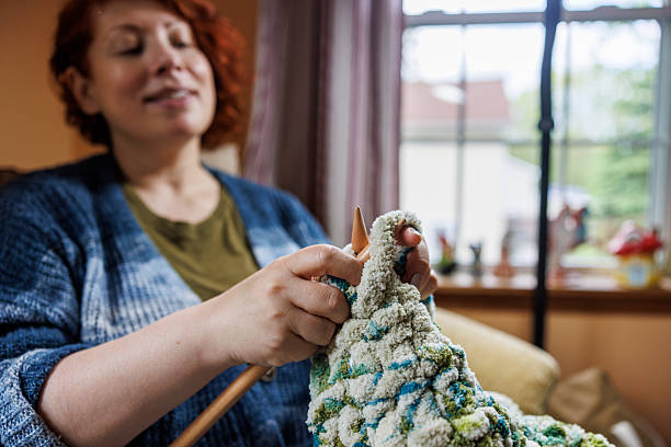 A 47-year-old Caucasian woman sits comfortably in her rural Pennsylvania home, working on a textured knitting project with traditional knitting needles. She holds light green and blue colored fabric with an intricate pattern, focusing on her handwork during a peaceful afternoon. Natural light streams through a multi-paned window behind her, illuminating the domestic interior setting. The scene captures a quiet moment of traditional fiber arts practiced as a relaxing hobby. The woman appears content and engaged with her needlework, demonstrating the meditative quality of this timeless craft.