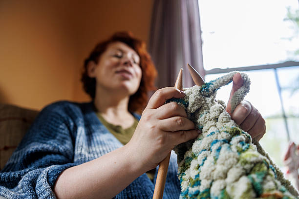 A 47-year-old Caucasian woman sits by a window working on a knitting project with wooden needles. She holds a colorful blanket or throw with blue, green, and cream colored yarn in a textured stitch pattern. The woman has curly dark hair and wears glasses while concentrating on her needlework. Natural light from the window illuminates her hobby workspace in what appears to be a rural Pennsylvania home setting. The scene captures a quiet moment of traditional fiber arts and handcraft skills.