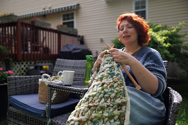 A 47-year-old Caucasian woman with curly red hair sits on an outdoor deck knitting a green and white patterned sweater. She holds wooden knitting needles and works with chunky yarn while seated in a comfortable chair. The rural Pennsylvania setting includes a residential deck with patio furniture, coffee cup, and wicker basket nearby. Lush greenery and residential buildings create a peaceful backyard environment for this traditional fiber arts hobby during daytime hours.