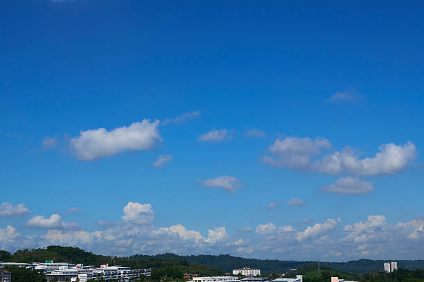 Scenic landscape featuring a vibrant blue sky dotted with fluffy white clouds, overlooking rolling green hills and modern buildings