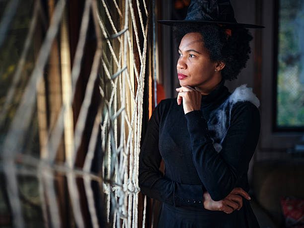 Pensive African American woman dressed up as a witch on Halloween looking through the window at home.