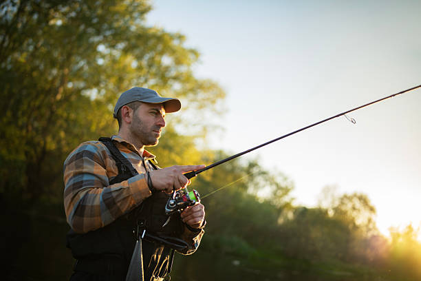 A mid-aged fisherman fly fishes in a tranquil river at sunset bathed by the warm glow of the fading sun.