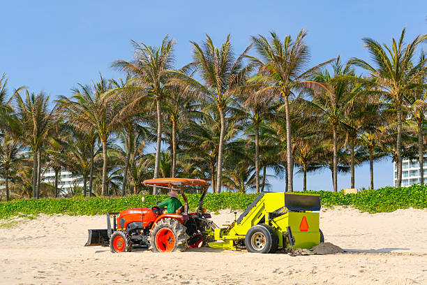 A red tractor with a screening unit trailer cleans sand from small debris in a beach area.