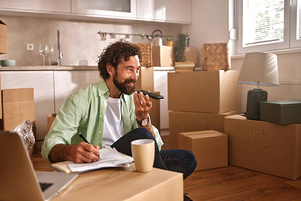 istock Man organizing a new home while speaking on the phone and taking notes in a cozy living space filled with unpacked boxes 2218196819