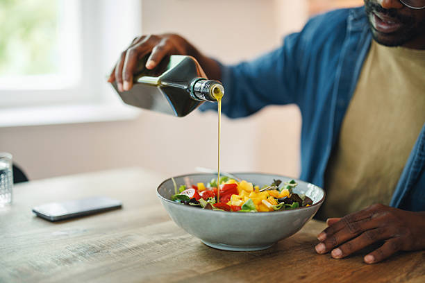 A Black man pours olive oil over a fresh salad in a home setting. The image conveys themes of self-care, healthy living, and relaxation, highlighting a focus on nutrition and personal well-being.