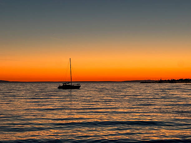A sailboat is gliding peacefully on the calm sea as a vibrant sunset is illuminating the sky with warm hues in Novalja. The serene water is reflecting the orange glow, creating a tranquil ambiance.