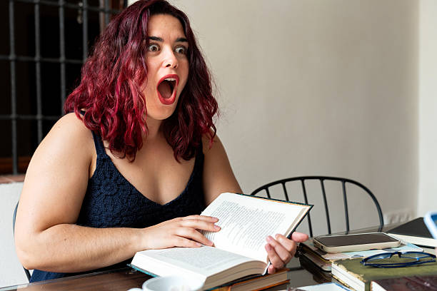 Redhead writer gasping while reading surprising plot twist in book at home office desk, surrounded by notes, mobile phone and glasses