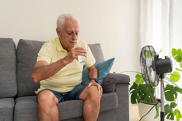 istock Senior man drinking water on sofa with electric fan cooling him down during summer heatwave at home 2218211635