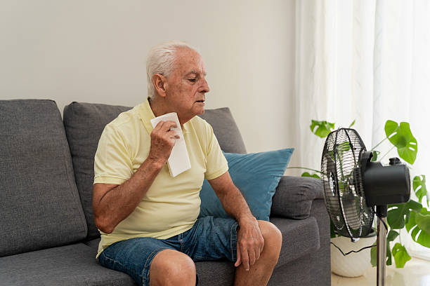istock Senior man sitting on sofa wiping sweat, feeling hot at home using electric fan 2218211639
