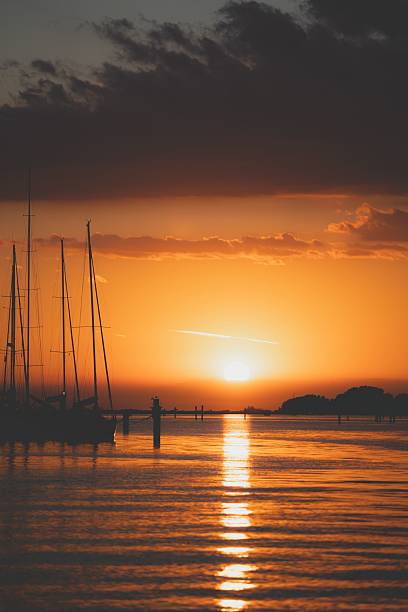 A serene sunset over a calm sea with silhouetted sailboats and a vibrant orange sky reflecting on the water.