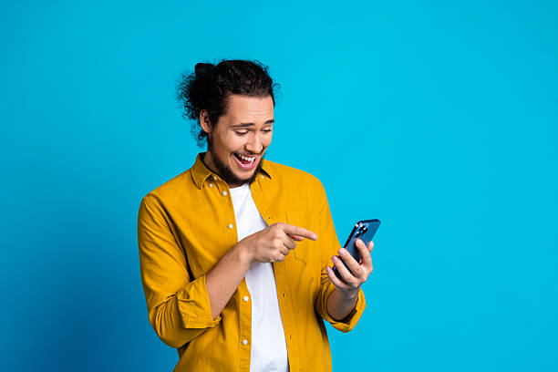 istock Young smiling man in yellow shirt using smartphone on blue background, browsing and interacting for leisure 2218223049
