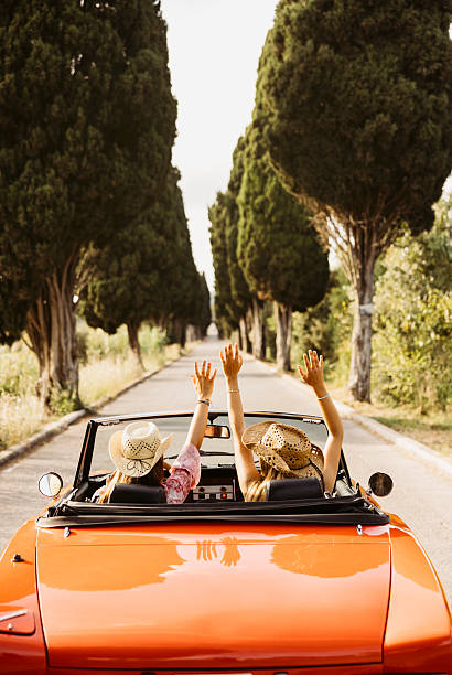 Two young women raising arms while driving orange convertible car on a country road