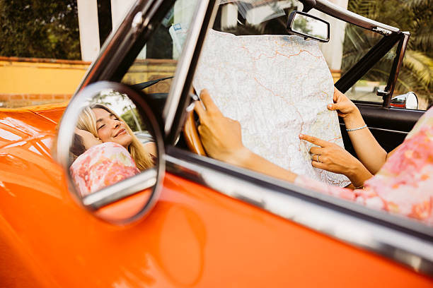 Two young women are looking at a map while sitting in a convertible car, planning their road trip