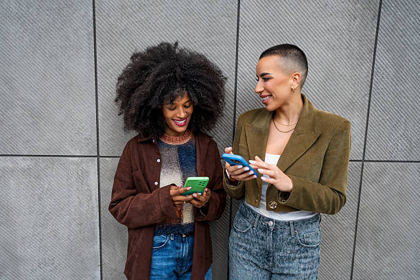 istock Two young women using smartphones and smiling by a gray wall 2218238460