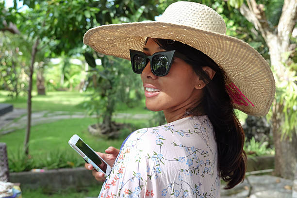 Close-up shot of an Indonesian woman wearing a sunhat and sunglasses holding a smartphone and smiling while standing in a sunny park or garden.\nconcept: summer fashion, happy woman, outdoor lifestyle, mobile phone use, digital connection, tropical vacation, stylish woman, nature background, travel influencer, casual elegance.