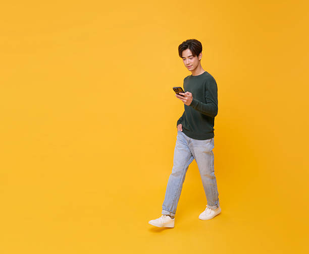istock Young smiling happy asian man 20s using mobile phone and walking isolated on yellow background. portrait People lifestyle concept 2218262409