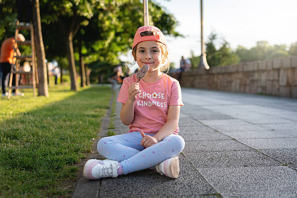 A smiling young girl sits cross-legged while eating ice cream in a sunny park. She wears a pink shirt with gentle lighting emphasizing the casual and joyful outdoor moment.