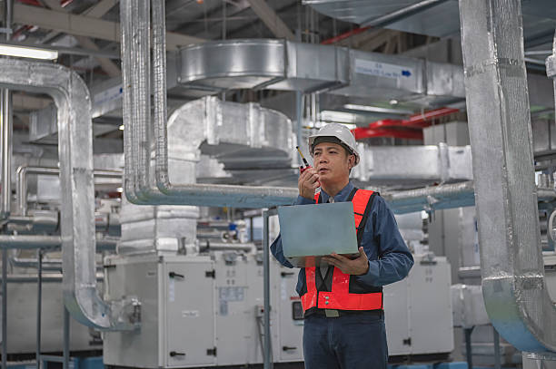 Service Engineer checking on Air handing unit room  and in hand hold computer laptop with  radio communication. He is wearing a white safety helmet and a high-visibility jacket, and appears to be checking data on a Laptop.