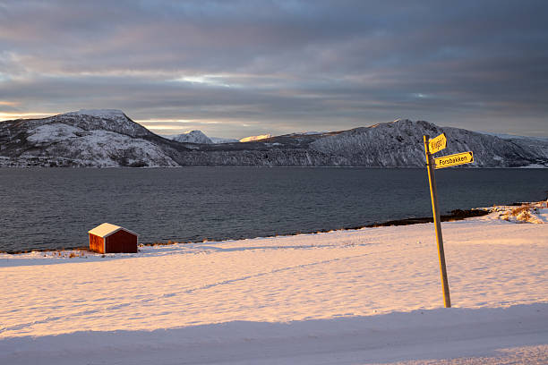 Sunrise in a cold day in the late arctic autumn. Nature covered by snow. Mountains on the horizon. Red cottage on the coast. Cloudy colorful sky. Lundenes, island Grytoya, Norway.