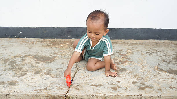A toddler playing with a red toy motorcycle on a sandy courtyard floor, engaging in solo imaginative play.