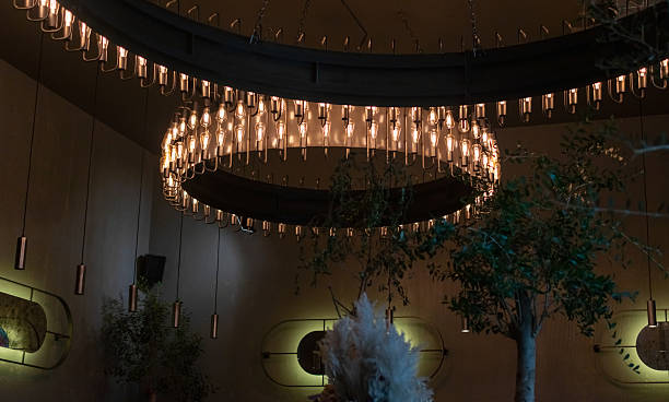 Dimly lit interior features two large circular chandeliers with vintage-style bulbs, surrounded by hanging pendant lights, decorative wall art, and indoor trees creating cozy, modern ambiance