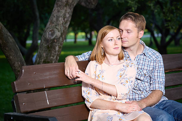 Beautiful young couple sitting together on the bench in the park. They are embracing and holding hands. Shooting in the summer in the evening