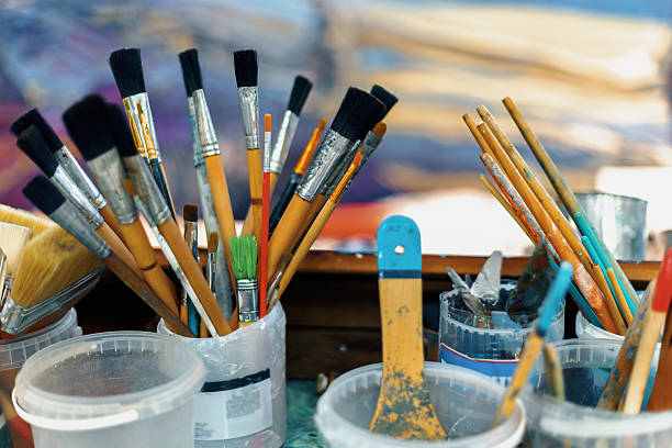 Many used paintbrushes and palette knives are seen resting in plastic containers in an artist's studio, with a colorful painting visible in the background