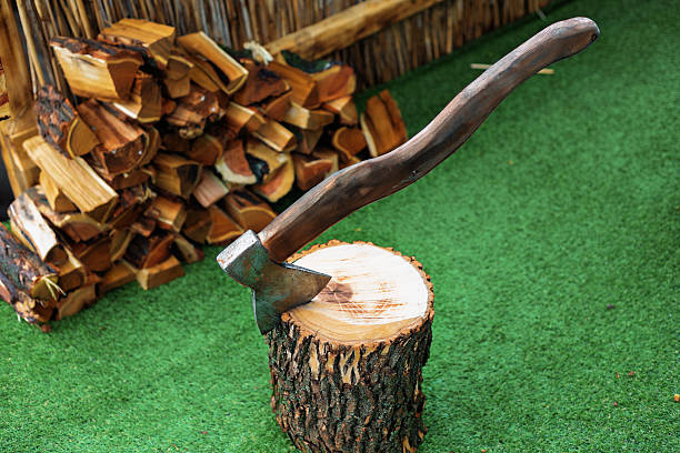 A tool for chopping wood rests on a wooden log covered in snow. Nearby, piles of chopped wood are stacked. A red cover is seen in the background suggesting outdoor storage.